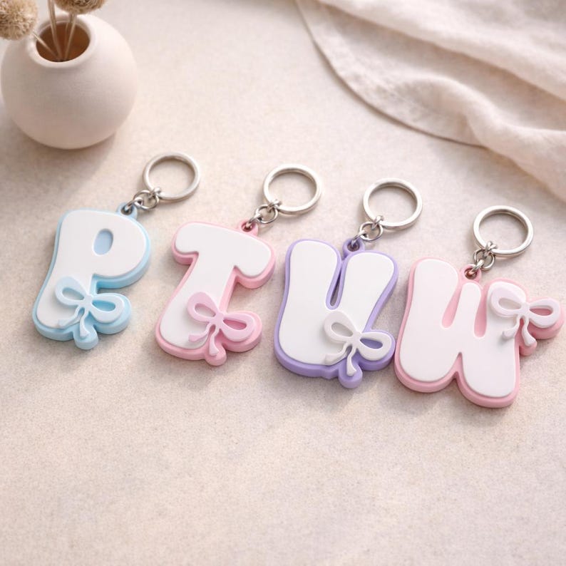 May include: Four letter-shaped keychains featuring the letters P, T, U, and W. Each keychain has a white center, a colored border in light blue, pink, lavender, or light pink, and a small matching bow detail attached to the side with a metal ring.
