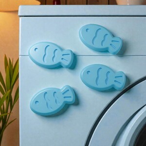 May include: Four light blue fish-shaped magnets are attached to a light blue surface. The magnets have a smooth, glossy finish and are arranged in a playful pattern. The fish magnets are a decorative item.