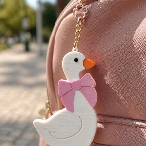 May include: A white goose keychain with an orange beak and feet, wearing a pink bow around its neck. The goose is attached to a gold-colored metal chain featuring a small flower-shaped link, which is clipped onto a dusty pink backpack zipper.