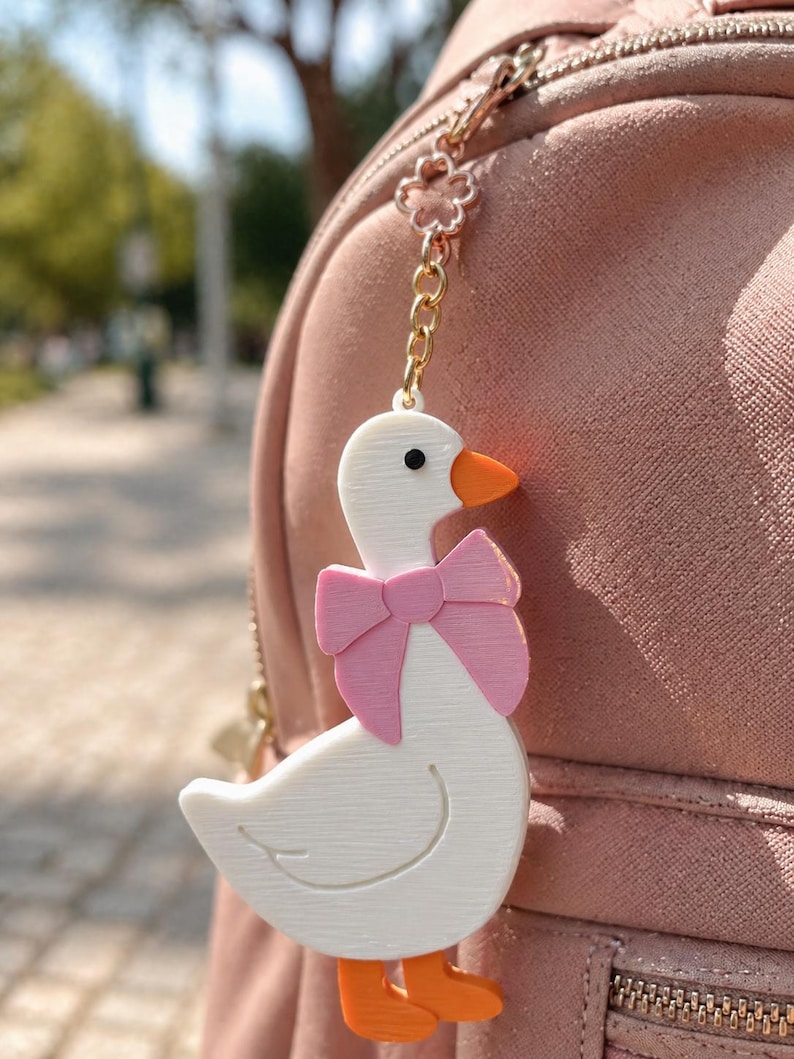 May include: A white goose keychain with an orange beak and feet, wearing a pink bow around its neck. The goose is attached to a gold-colored metal chain featuring a small flower-shaped link, which is clipped onto a dusty pink backpack zipper.