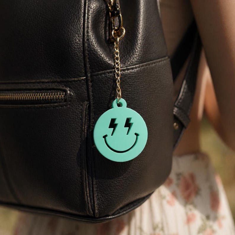 K&ouml;nnte beinhalten: Ein runder Schl&uuml;sselanh&auml;nger aus mintgr&uuml;nem Acryl mit einem ausgeschnittenen Smiley-Gesicht-Design und Augen in Blitzform. Der Anh&auml;nger ist an einer goldfarbenen Metallkette mit Karabinerverschluss befestigt, die an einem schwarzen Lederrucksack h&auml;ngt.