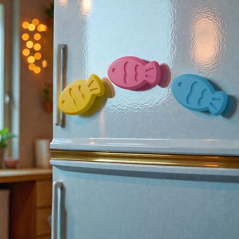 May include: Three colorful fish-shaped magnets are attached to a light blue refrigerator door. The magnets are yellow, pink, and blue. The refrigerator has a gold-colored trim and a silver handle. The background is slightly blurred.