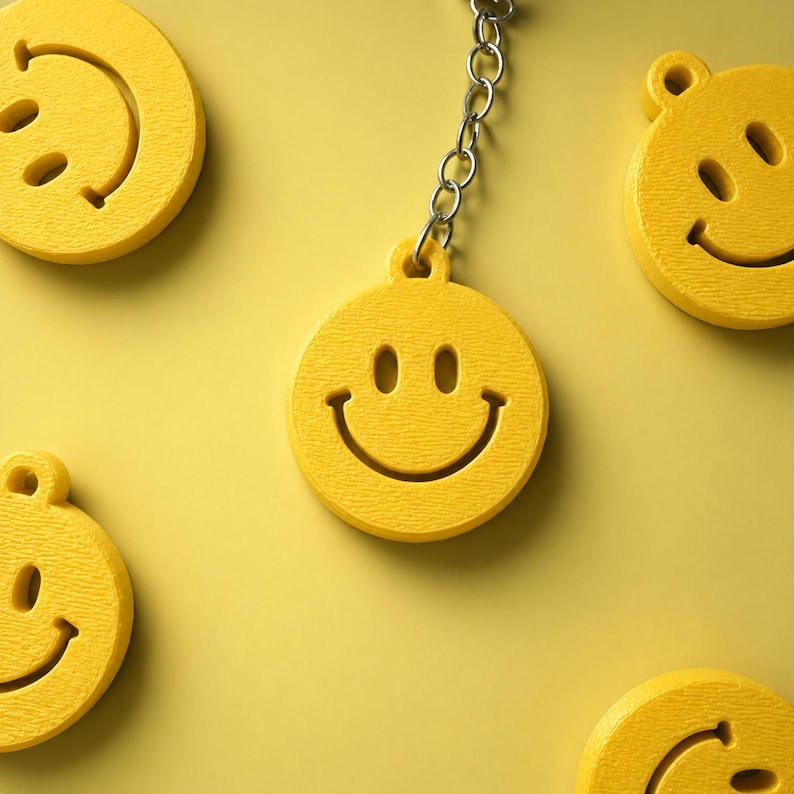 K&ouml;nnte beinhalten: Ein hellgelber, kreisf&ouml;rmiger Schl&uuml;sselanh&auml;nger mit einem ausgeschnittenen Smiley-Gesicht, bestehend aus zwei ovalen Augen und einem geschwungenen Mund. Der Anh&auml;nger ist an einer silbernen Metallkette befestigt, umgeben von vier weiteren gelben Smileys.