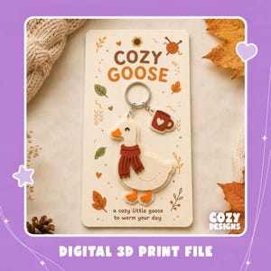 Könnte beinhalten: Ein weißer Gänse-Schlüsselanhänger mit orangefarbenem Schnabel und Füßen, der einen dunkelorangen Schal trägt, befestigt an einem silbernen Ring mit einem kleinen dunkelorangen Tassenanhänger mit weißem Herz. Die Karte zeigt den Text COZY GOOSE und a cozy little goose to warm your day.