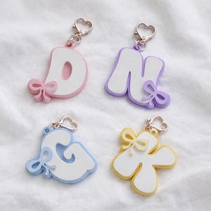 May include: Four letter keychains featuring the letters D, N, G, and K. Each letter is white with a colored border in pink, purple, light blue, or yellow. Each keychain includes a small matching bow accent and a silver heart-shaped lobster clasp.