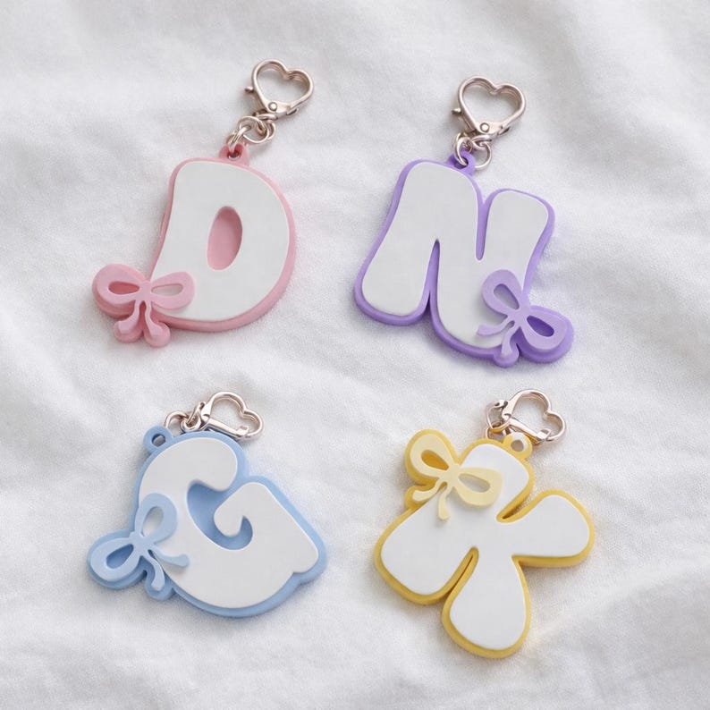 May include: Four letter keychains featuring the letters D, N, G, and K. Each letter is white with a colored border in pink, purple, light blue, or yellow. Each keychain includes a small matching bow accent and a silver heart-shaped lobster clasp.