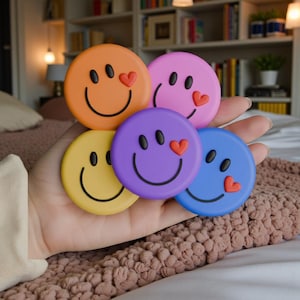 以下が含まれることがあります： 手には、5つのカラフルな丸いスマイリーフェイスマグネットがあります。各マグネットには、黒い笑顔、2つの黒い目、赤いハートがあります。マグネットはオレンジ、ピンク、紫、青、黄色です。