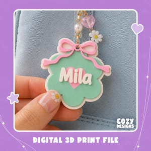 Puede incluir: Un llavero de acrílico personalizado con forma de flor verde menta, un corazón rosa y el nombre Mila en letras blancas. El dije tiene un lazo rosa y está unido a una cadena dorada con una perla blanca, un corazón rosa y una margarita blanca.