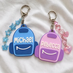 Puede incluir: Dos llaveros con forma de mochila y anillas met&aacute;licas. El azul lleva el nombre Michael y un colgante de estrella azul brillante. El rosa lleva el nombre Rebecca y un colgante de coraz&oacute;n rosa. Ambos tienen una l&iacute;nea blanca curva al frente.