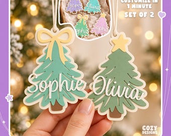 May include: Two layered Christmas tree ornaments with white string hangers. One tree features a yellow bow and the name Sophie, the other a yellow star and the name Olivia. Text reads: Customize in 1 minute, set of 2, and digital 3D print file.