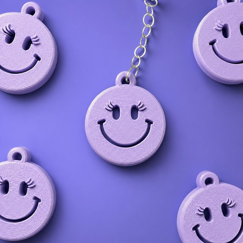 K&ouml;nnte beinhalten: Eine Sammlung hellvioletter, kreisf&ouml;rmiger Anh&auml;nger mit einem ausgeschnittenen Smiley-Gesicht und Wimpern. Jeder Anh&auml;nger hat oben eine kleine &Ouml;se, und ein Anh&auml;nger ist an einer silberfarbenen Metallkette vor einem violetten Hintergrund befestigt.