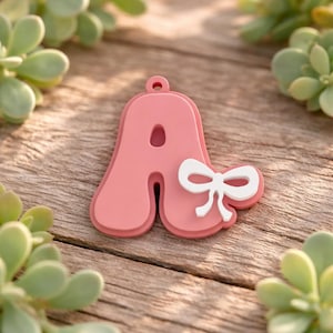 May include: A pink letter A charm with a smooth, rounded font and a small hole at the top for hanging. A solid white bow is attached to the right side of the letter. The charm has a layered, dimensional appearance with a matte finish.