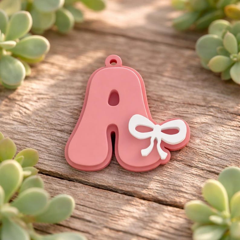 May include: A pink letter A charm with a smooth, rounded font and a small hole at the top for hanging. A solid white bow is attached to the right side of the letter. The charm has a layered, dimensional appearance with a matte finish.