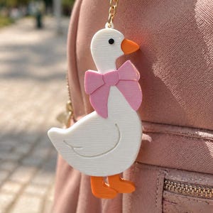 Op de afbeelding: Een witte ganzen sleutelhanger met een oranje snavel en poten, en een roze strik. De sleutelhanger hangt aan een roze rugzak. De gans heeft een textuur en een zwart oog.