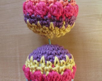 May include: Two handmade, crocheted spheres suspended by a blue string. The spheres feature a vibrant color palette of pink, yellow, purple, and blue. The crochet work includes textured, raised floral patterns, creating a decorative and tactile appeal.