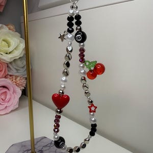 May include: A black and white beaded phone charm with a red heart, a red star, a black 8-ball, and a red cherry. The charm is hanging from a black string.