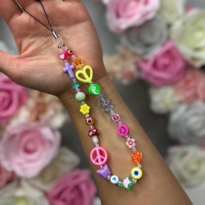May include: A colourful phone charm with a variety of beads, including peace signs, hearts, stars, smiley faces, and an evil eye. The charm is attached to a black string.