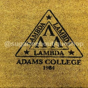 Puede incluir: Una alfombra de bienvenida dorada con un diseño de triángulo negro que incluye el texto "Lambda Adams College 1984".