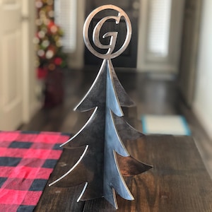 May include: A metal Christmas tree with a circular top featuring the letter "G". The tree has a layered design with multiple branches. It is displayed on a dark wooden surface with a red and black plaid table runner.