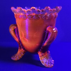May include: An orange glass footed vase with a ruffled rim and ornate handles. The vase has three feet and a textured surface. The glass has a glossy finish and is set against a blue background.