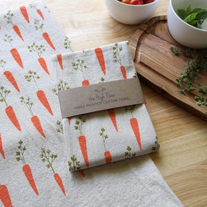 Puede incluir: Dos pa&ntilde;os de cocina de algod&oacute;n estampados a mano con un dise&ntilde;o de zanahorias. Las zanahorias son de color naranja con hojas verdes, impresas sobre un fondo beige claro. Uno de los pa&ntilde;os est&aacute; doblado y envuelto con una banda de papel marr&oacute;n que dice "The High Floor HAND PRINTED COTTON TOWEL".