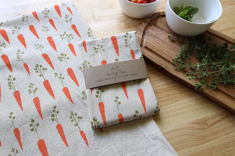 Puede incluir: Dos pa&ntilde;os de cocina de algod&oacute;n estampados a mano con un dise&ntilde;o de zanahorias. Las zanahorias son de color naranja con hojas verdes, impresas sobre un fondo beige claro. Uno de los pa&ntilde;os est&aacute; doblado y envuelto con una banda de papel marr&oacute;n que dice "The High Floor HAND PRINTED COTTON TOWEL".
