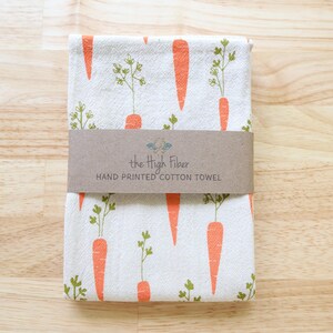 Puede incluir: Toalla de algod&oacute;n blanca doblada con un patr&oacute;n repetido de zanahorias naranjas y hojas verdes. Una banda de papel marr&oacute;n envuelve la toalla, con el texto "the High Fiber HAND PRINTED COTTON TOWEL".