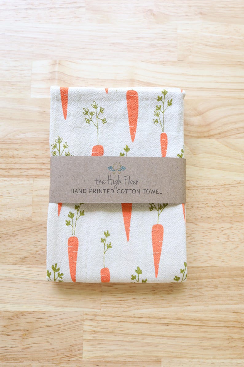 Puede incluir: Toalla de algod&oacute;n blanca doblada con un patr&oacute;n repetido de zanahorias naranjas y hojas verdes. Una banda de papel marr&oacute;n envuelve la toalla, con el texto "the High Fiber HAND PRINTED COTTON TOWEL".