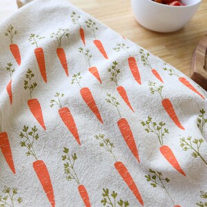 Puede incluir: Un pa&ntilde;o de cocina beige con un patr&oacute;n repetido de zanahorias naranjas y hojas verdes. El pa&ntilde;o est&aacute; doblado, mostrando el patr&oacute;n completo. El fondo incluye una superficie de madera y un cuenco blanco.