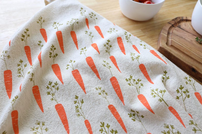Puede incluir: Un pa&ntilde;o de cocina beige con un patr&oacute;n repetido de zanahorias naranjas y hojas verdes. El pa&ntilde;o est&aacute; doblado, mostrando el patr&oacute;n completo. El fondo incluye una superficie de madera y un cuenco blanco.