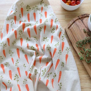 Puede incluir: Un pa&ntilde;o de cocina blanco con un patr&oacute;n repetido de zanahorias naranjas y hojas verdes. El pa&ntilde;o est&aacute; colocado sobre una superficie de madera, con cuencos de tomates y albahaca cerca. Tambi&eacute;n se ven una tabla de cortar de madera y ramitas de tomillo.