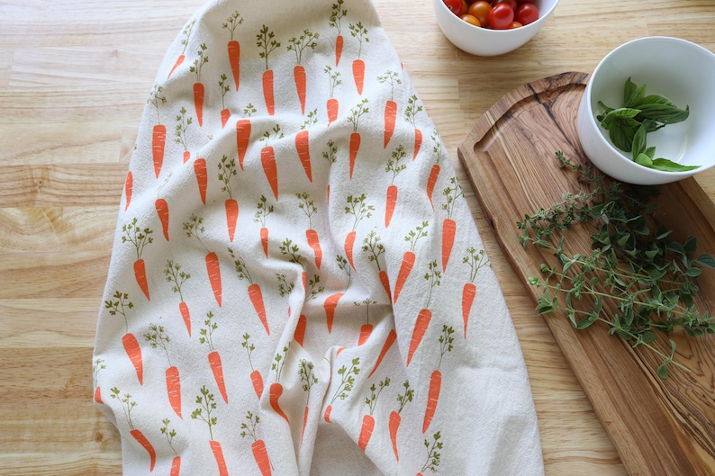 Puede incluir: Un pa&ntilde;o de cocina blanco con un patr&oacute;n repetido de zanahorias naranjas y hojas verdes. El pa&ntilde;o est&aacute; colocado sobre una superficie de madera, con cuencos de tomates y albahaca cerca. Tambi&eacute;n se ven una tabla de cortar de madera y ramitas de tomillo.