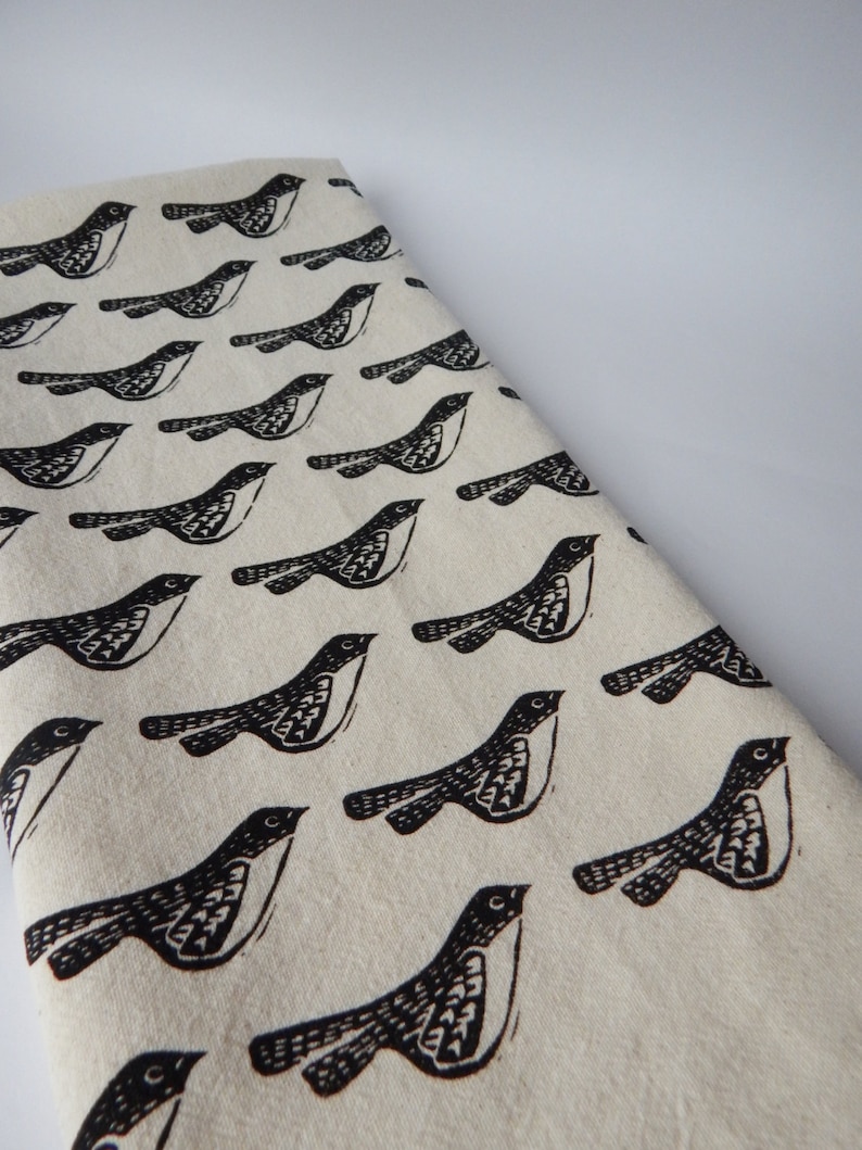 May include: A white fabric tea towel with a repeating pattern of black stylized birds. The birds are facing right and have a simple design.