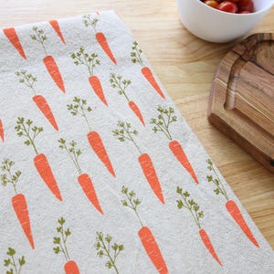 Puede incluir: Un pa&ntilde;o de cocina con un patr&oacute;n repetido de zanahorias naranjas con hojas verdes sobre un fondo beige claro. El pa&ntilde;o es parcialmente visible sobre una superficie de madera, con cuencos y una tabla de cortar en el fondo.