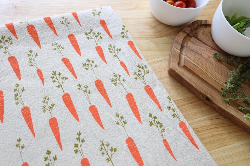 Puede incluir: Un pa&ntilde;o de cocina con un patr&oacute;n repetido de zanahorias naranjas con hojas verdes sobre un fondo beige claro. El pa&ntilde;o es parcialmente visible sobre una superficie de madera, con cuencos y una tabla de cortar en el fondo.