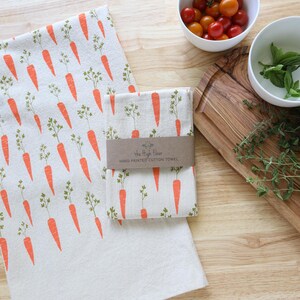 Puede incluir: Toallas de cocina de algod&oacute;n blanco con un patr&oacute;n repetido de zanahorias naranjas y hojas verdes. Una toalla est&aacute; doblada y envuelta con una banda de papel marr&oacute;n. Cerca hay cuencos con tomates y hierbas.