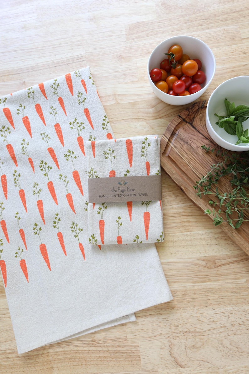 Puede incluir: Toallas de cocina de algod&oacute;n blanco con un patr&oacute;n repetido de zanahorias naranjas y hojas verdes. Una toalla est&aacute; doblada y envuelta con una banda de papel marr&oacute;n. Cerca hay cuencos con tomates y hierbas.