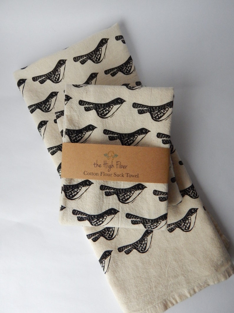May include: Two cream-colored cotton flour sack towels with a black bird print. The towels are folded and stacked on top of each other. A brown paper tag with the text "the High Fiber Cotton Flour Sack Towel" is attached to the top towel.