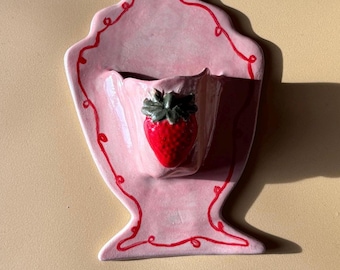 Może przedstawiać: Różowa ceramiczna kieszeń ścienna z akcentem truskawkowym. Kieszeń ma falisty brzeg z czerwonymi detalami. Truskawka jest czerwona z zielonymi liśćmi. Kieszeń ścienna jest przeznaczona do przechowywania małych roślin lub przedmiotów dekoracyjnych.