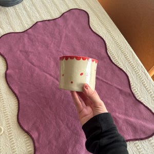 May include: A small, cylindrical ceramic pot with a cream-colored base and red accents. The rim is scalloped and painted red, with red dots scattered across the surface. The pot is held over a purple placemat.