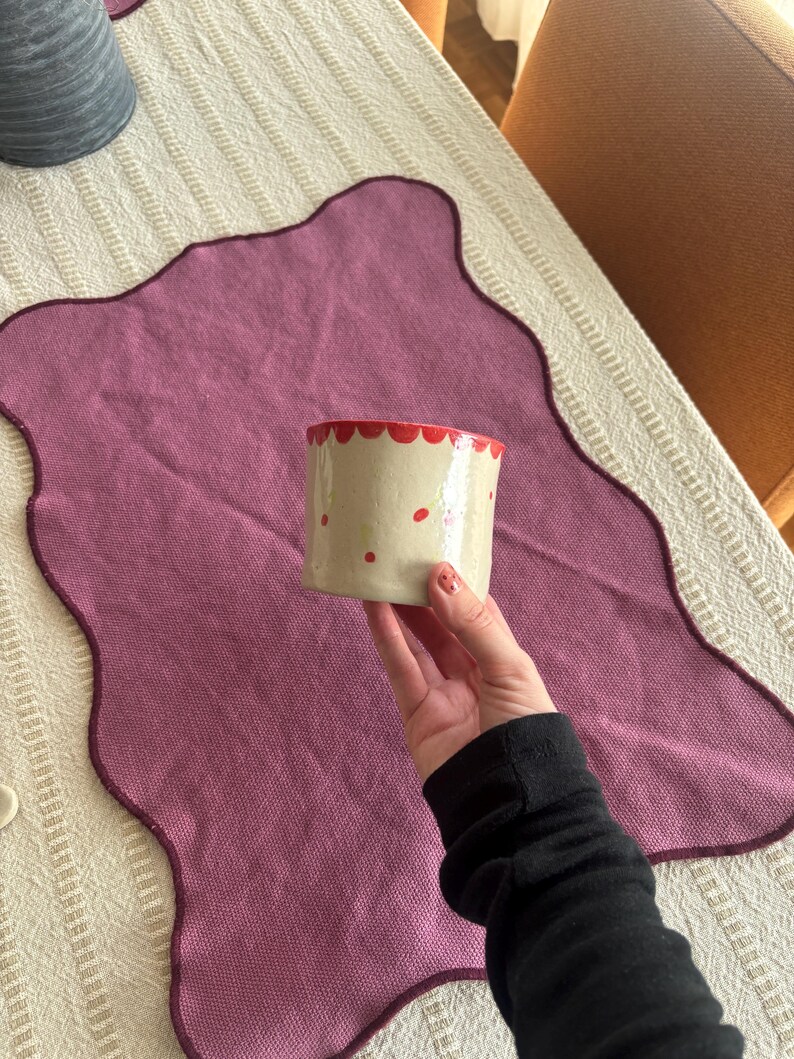 May include: A small, cylindrical ceramic pot with a cream-colored base and red accents. The rim is scalloped and painted red, with red dots scattered across the surface. The pot is held over a purple placemat.
