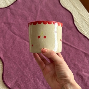 May include: A cylindrical ceramic mug with a cream-colored base, adorned with red cherries and a scalloped red rim. The mug is held against a purple background, showcasing its handmade design and playful aesthetic.