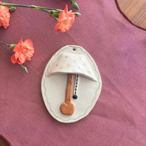 May include: A white ceramic wall hanging shaped like a lamp with a floral pattern on the lampshade. The lamp has a brown base and a black pull chain.