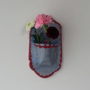 May include: A blue and red ceramic wall pocket with a scalloped edge, holding a small bouquet of flowers. The flowers include a large pink carnation, a dark red flower, and a white and green flower.