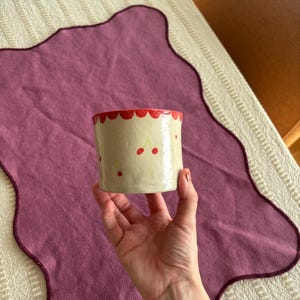 May include: A cylindrical ceramic cup with a cream-colored base, adorned with red cherries and a scalloped red rim. The cup is held in a hand, showcasing its handmade design. The cup is approximately 3 inches tall.