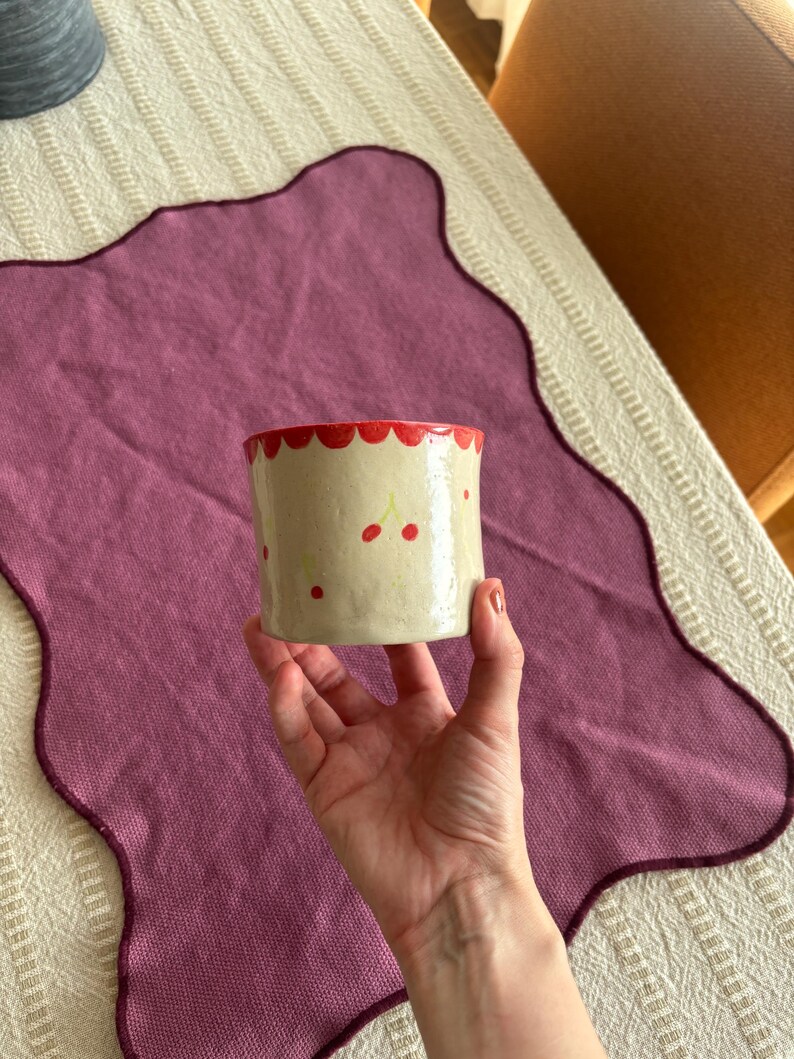 May include: A cylindrical ceramic cup with a cream-colored base, adorned with red cherries and a scalloped red rim. The cup is held in a hand, showcasing its handmade design. The cup is approximately 3 inches tall.