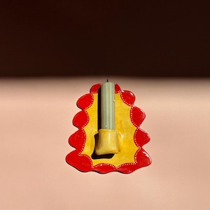 May include: A handmade ceramic candle holder with a yellow base and a red scalloped border. The holder has a small yellow cup for the candle and holds a light green candle. The background is pink.