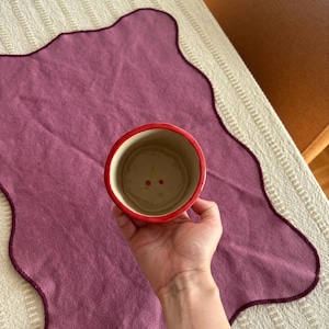 May include: A red and cream-colored ceramic cup with a cherry design inside. The cup is held over a purple placemat with a scalloped edge. The placemat is on a cream-colored tablecloth.