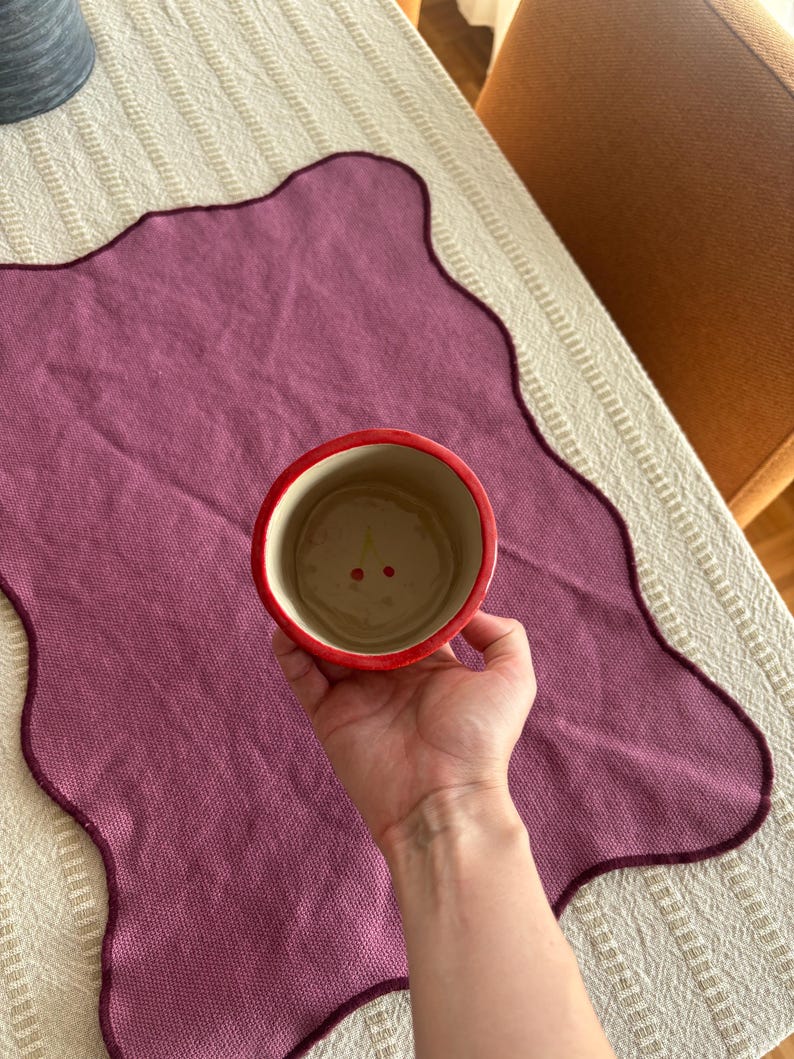 May include: A red and cream-colored ceramic cup with a cherry design inside. The cup is held over a purple placemat with a scalloped edge. The placemat is on a cream-colored tablecloth.