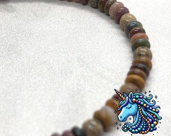 May include: A multi-colored beaded bracelet with a variety of brown, pink, and green stones. The bracelet is made with a mix of round and oval-shaped beads. The bracelet is accented with a single white bead. The bracelet is made by Unicorn Beading Company.
